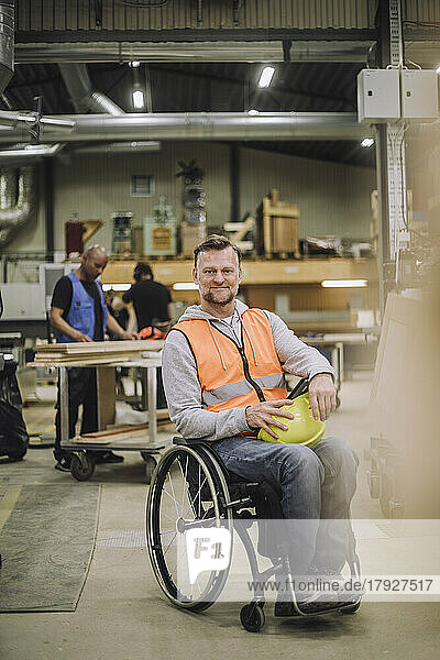 Portrait of smiling carpenter sitting on wheelchair with colleagues working in background at warehouse