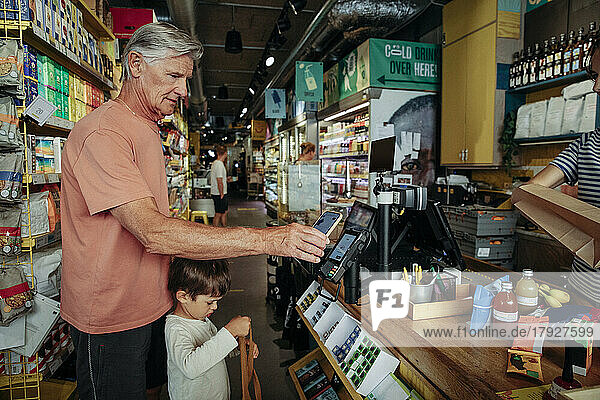 Seitenansicht eines älteren Mannes  der online bezahlt  während er mit seinem Enkel an der Kasse eines Supermarktes steht