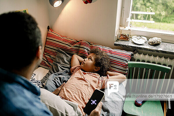 Junge schaut auf eine beleuchtete elektrische Lampe  während sein Vater sein Smartphone auf dem Bett zu Hause hält