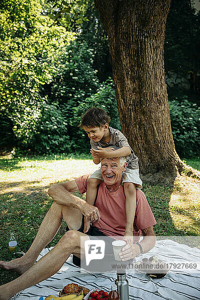 Großvater trägt seinen Enkel auf der Schulter  während er auf einer Picknickdecke im Park sitzt