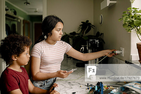 Mädchen lädt ihr Smartphone auf  während sie mit ihrem Bruder in der Nähe der Küchentheke zu Hause steht