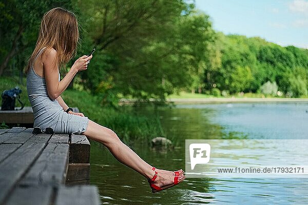 Seitenansicht eines Mädchens  das auf einem Holzsteg am Flussufer sitzt und Nachrichten schreibt. Schöne Frau mit Haar mit dem Wind geblasen