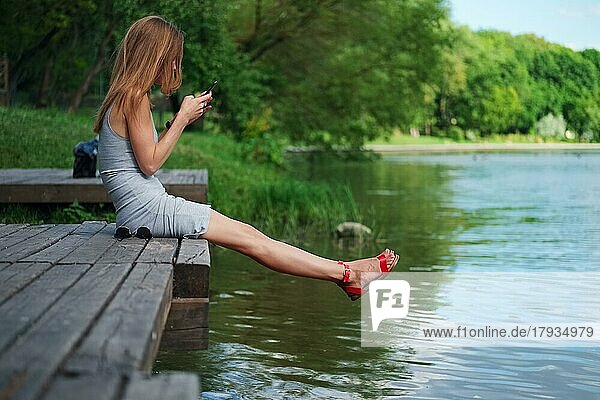 Seitenansicht eines Mädchens  das auf einem Holzsteg am Flussufer sitzt und Nachrichten schreibt. Schöne Frau mit Haar mit dem Wind geblasen