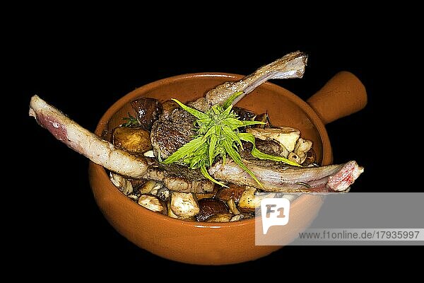 Drei gegrillte Lammkotelettes auf gebratenen Egerlinge (Agaricus) (Syn. : Psalliota)  in einer spanischen feuerfesten Keramikschüssel (Cazuela)  Foodfotografie mit schwarzem Hintergrund