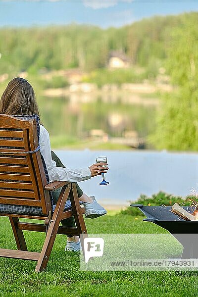 Junge Frau mit einem Glas Sangria in der Hand entspannt sich in der Natur neben einem Grill sitzend  Rückansicht