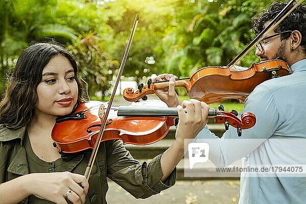 Geiger Mann und Frau Rücken an Rücken spielen Geige in einem Park im Freien. Zwei junge Geiger stehen und spielen Geige in einem Park. Porträt von Mann und Frau zusammen spielen Geige im Park
