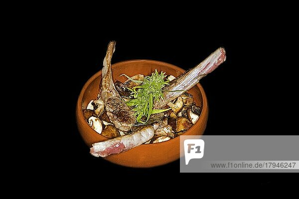 Drei gegrillte Lammkotelettes auf gebratenen Egerlinge (Agaricus) (Syn. : Psalliota)  in einer spanischen feuerfesten Keramikschüssel (Cazuela)  Foodfotografie mit schwarzem Hintergrund