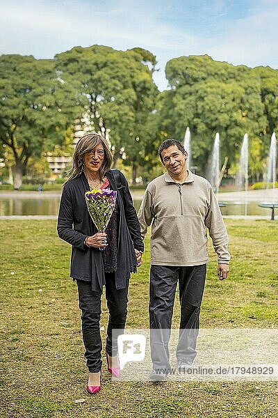 Reife Transgender Frau  die Hand in Hand mit ihrem Freund in einem Park spazieren geht und sich freut  weil er ihr Blumen geschenkt hat