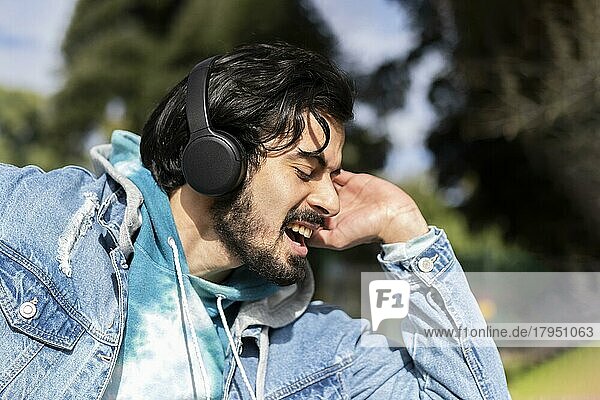Junger lateinamerikanischer Mann  der im Freien mit Kopfhörern Musik hört. Ausdruck von Glück  gewinnende Haltung