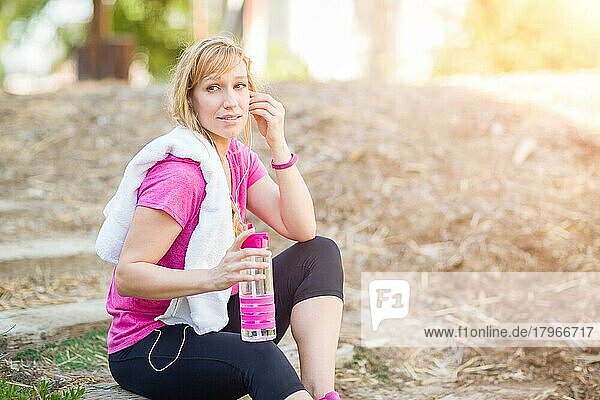 Junge fitte erwachsene Frau im Freien mit Handtuch und Wasserflasche in Trainingskleidung  die mit Kopfhörern Musik hört