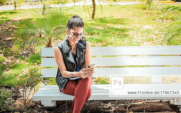 Mädchen sitzt auf einer Bank und überprüft ihr Handy  Glückliche Frau sitzt in einem Park und schreibt eine SMS auf ihrem Handy  Frau sitzt auf einer Bank und sendet eine SMS
