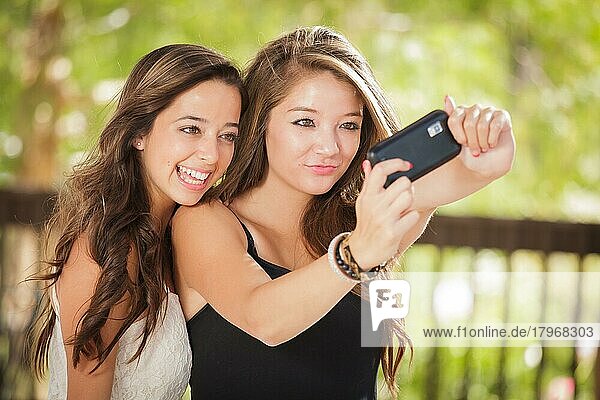 Zwei attraktive gemischtrassige Freundinnen benutzen ihre Smartphones für ein Selfie im Freien