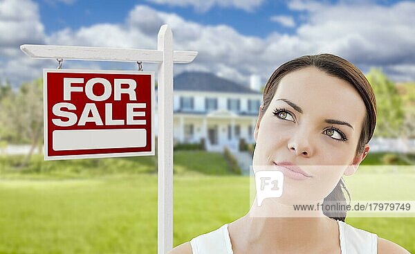 Nachdenkliche hübsche gemischtrassige Frau vor Haus und Haus zu verkaufen Immobilien Schild nach oben und zur Seite schauen