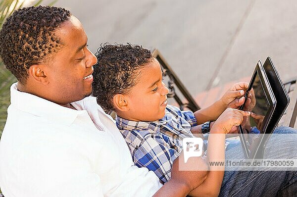 Afroamerikanischer Vater und gemischtrassiger Sohn verwenden Computer-Tablet auf einer Bank im Park