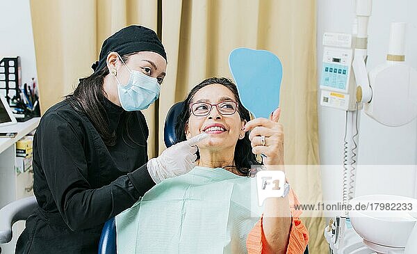 Zufriedene weibliche Klientin in der Zahnklinik  die in den Spiegel schaut  Zahnarzt mit Patientin  die in der Praxis in den Handspiegel lächelt  weibliche Patientin  die die Zähne nach der Zahnbehandlung in der Zahnklinik kontrolliert