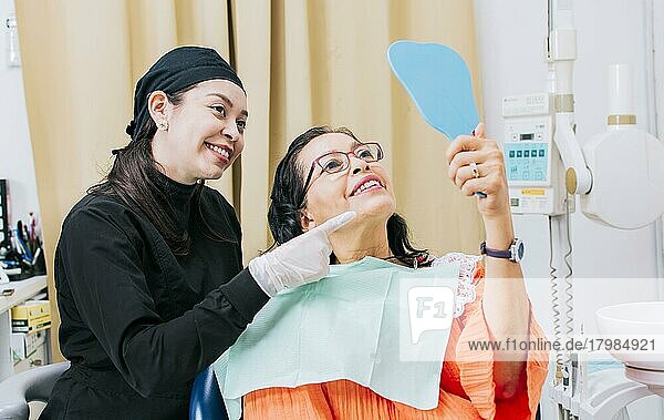 Satisfied female client in dental clinic looking at mirror  Dentist with patient smiling at hand mirror in office  female patient checking teeth after curing teeth in dental clinic