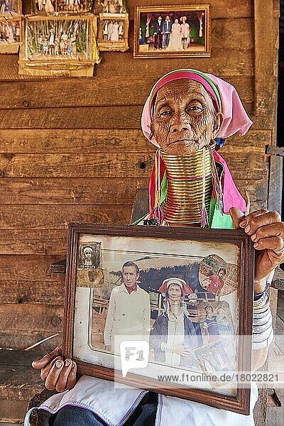 Ederly Kayan Lahwi Frau (80 Jahre alt) mit Messinghalsschleifen und traditioneller Kleidung  die auf ihrem Heimdeck sitzt und Bilder von ihr im Alter von 13 Jahren zeigt  als sie begann  die Schleifen zu tragen  Pan Pet Region  Kayah State  Myanmar  Asien