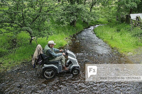 Älterer Mann mit Mobilitätsproblemen besucht mit dem Tramper (Geländescooter) Bauernhof  überquert Bach an Furt  Whitewell  Forest of Bowland  Lancashire  England  Juli