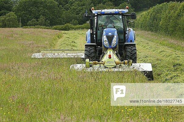Lohnunternehmer mit New Holland Traktor T6080 mit front- und heckmontierten Claas-Mähwerken  Mähen von Gras für Silage auf Biobetrieb  Powys  Wales  Juni