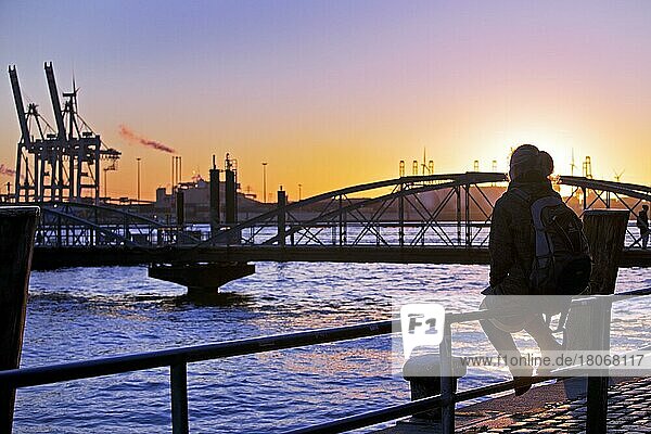 Eine Frau sitzt auf einem Geländer bei Sonnenuntergang an der Norderelbe in Altona  Hamburger Hafen  Hamburg  Deutschland  Europa