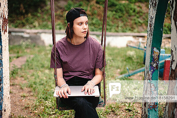 Junge Frau mit Laptop auf einer Schaukel auf einem verlassenen Spielplatz sitzend