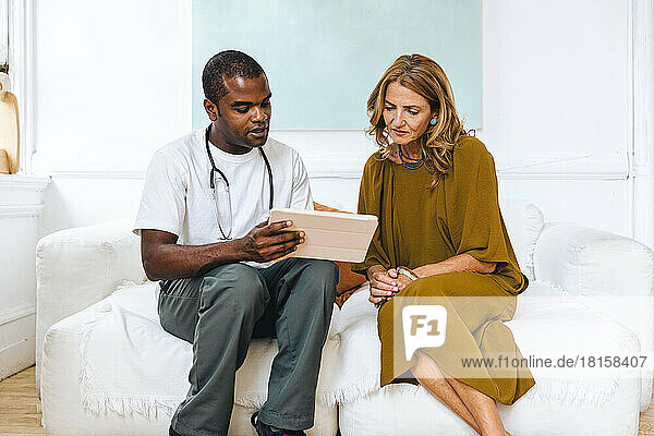 Arzt im Gespräch mit einem Patienten über einen Tablet-PC  während er auf dem Sofa sitzt