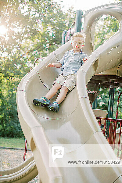 Ein blonder 5-jähriger Junge rutscht bei Sonnenuntergang eine Spielplatzrutsche hinunter.