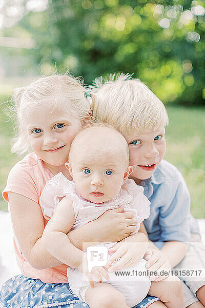 Two blonde toddlers hug their blue-eyed baby sister in park.