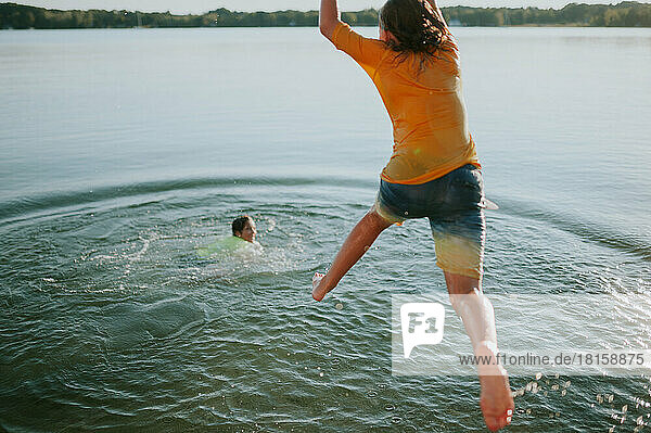 Junge springt vom Steg  während ein anderer Junge lächelnd im Wasser schwimmt