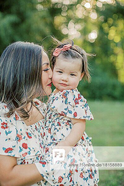 1-year-old girl in floral dress is held close by Asian mother.
