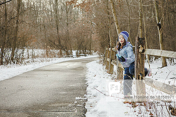 A little girl sits on wood fence along snowy fence with binoculars