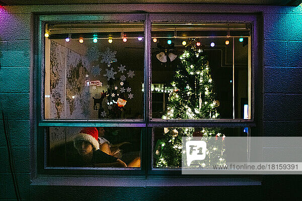 Junge schaut aus dem Fenster seines Hauses mit Weihnachtsbaum und Weihnachtsmannmütze
