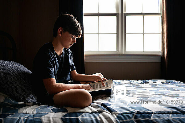 Young boy quietly reading a book on his bed in his bedroom.
