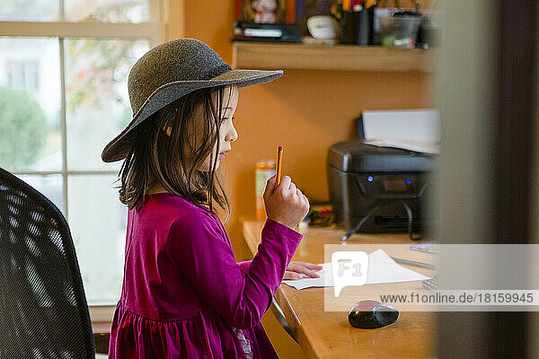 Ein kleines Kind mit Hut sitzt am Schreibtisch mit Papier und Bleistift