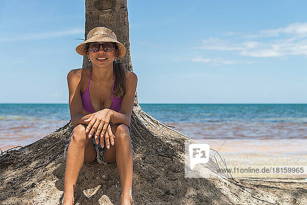 Lateinamerikanische Frau unter einer Palme am Strand sitzend und einen Hut tragend