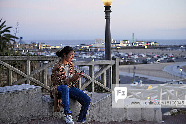 Woman using mobile phone while sitting on retaining wall at sunset