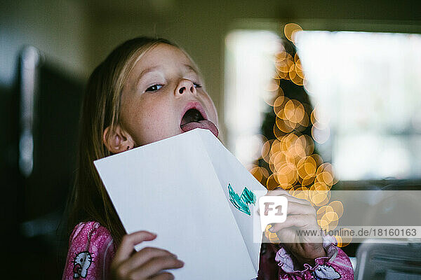 Girl licks letter for Santa in front of Christmas tree lights