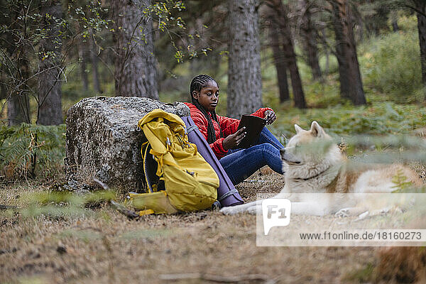 Young woman reading book sitting with dog in forest