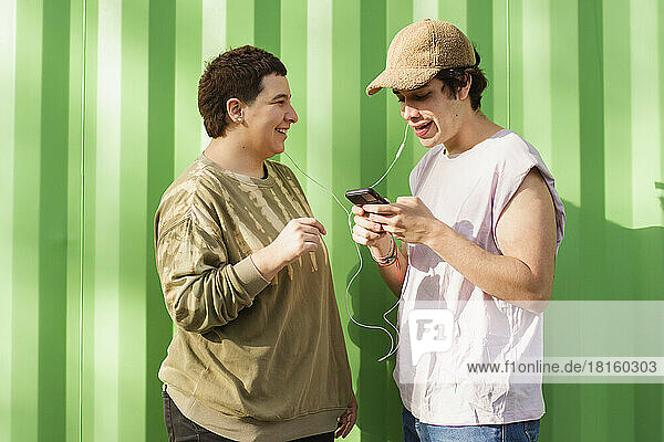 Happy non-binary couple using smart phone and listening to music with In-ear headphones