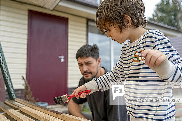 Father looking at son painting wooden swing in front of house