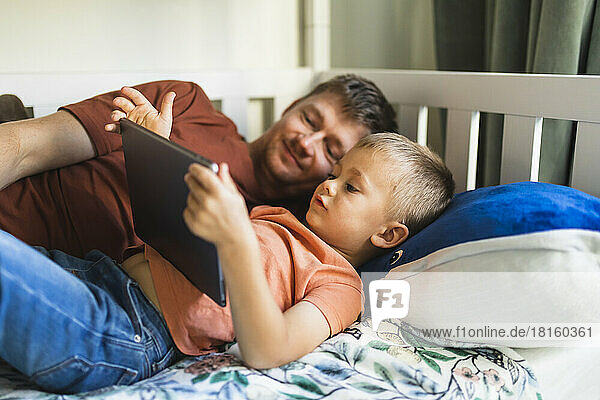 Son and father watching tablet PC lying on bed at home
