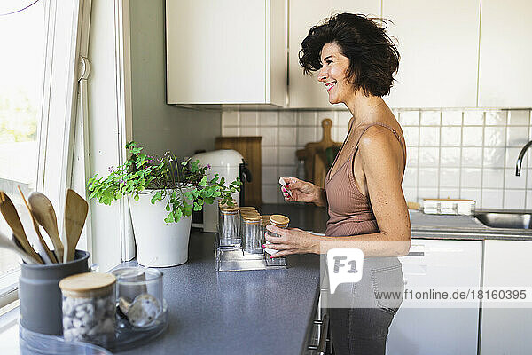 Smiling woman holding jar looking through window standing at kitchen counter