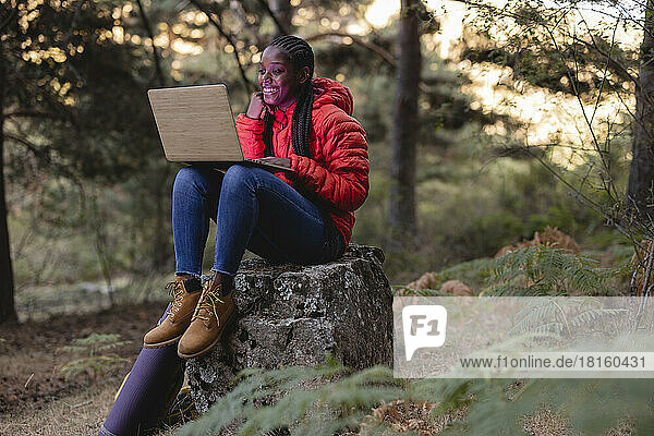 Happy young woman using laptop sitting on rock in forest