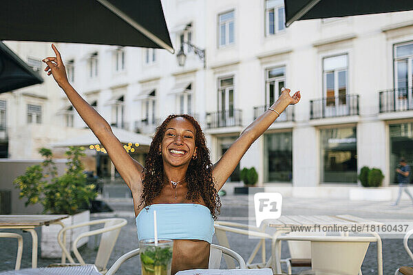 Happy young woman with arms raised sitting at sidewalk cafe