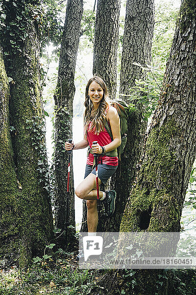 Smiling woman with hiking poles leaning on tree trunk