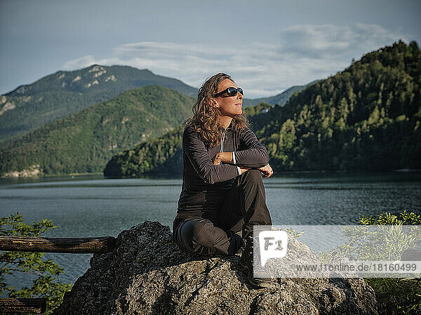Smiling mature woman wearing sunglasses sitting on rock in front of lake