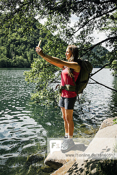 Hiker taking selfie through smart phone standing on rock near Lake Levico