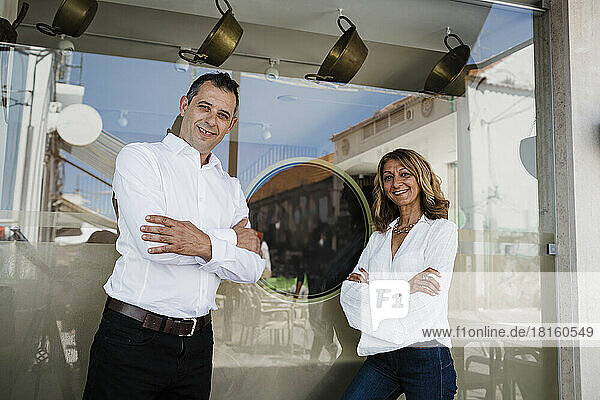 Happy restaurant owners with arms crossed standing by glass window