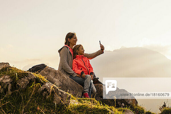 Happy mother with daughter taking selfie through mobile phone sitting on rock