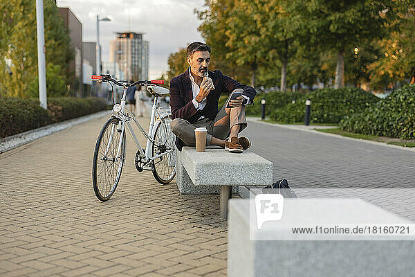 Businessman having lunch sitting on bench by bicycle at footpath